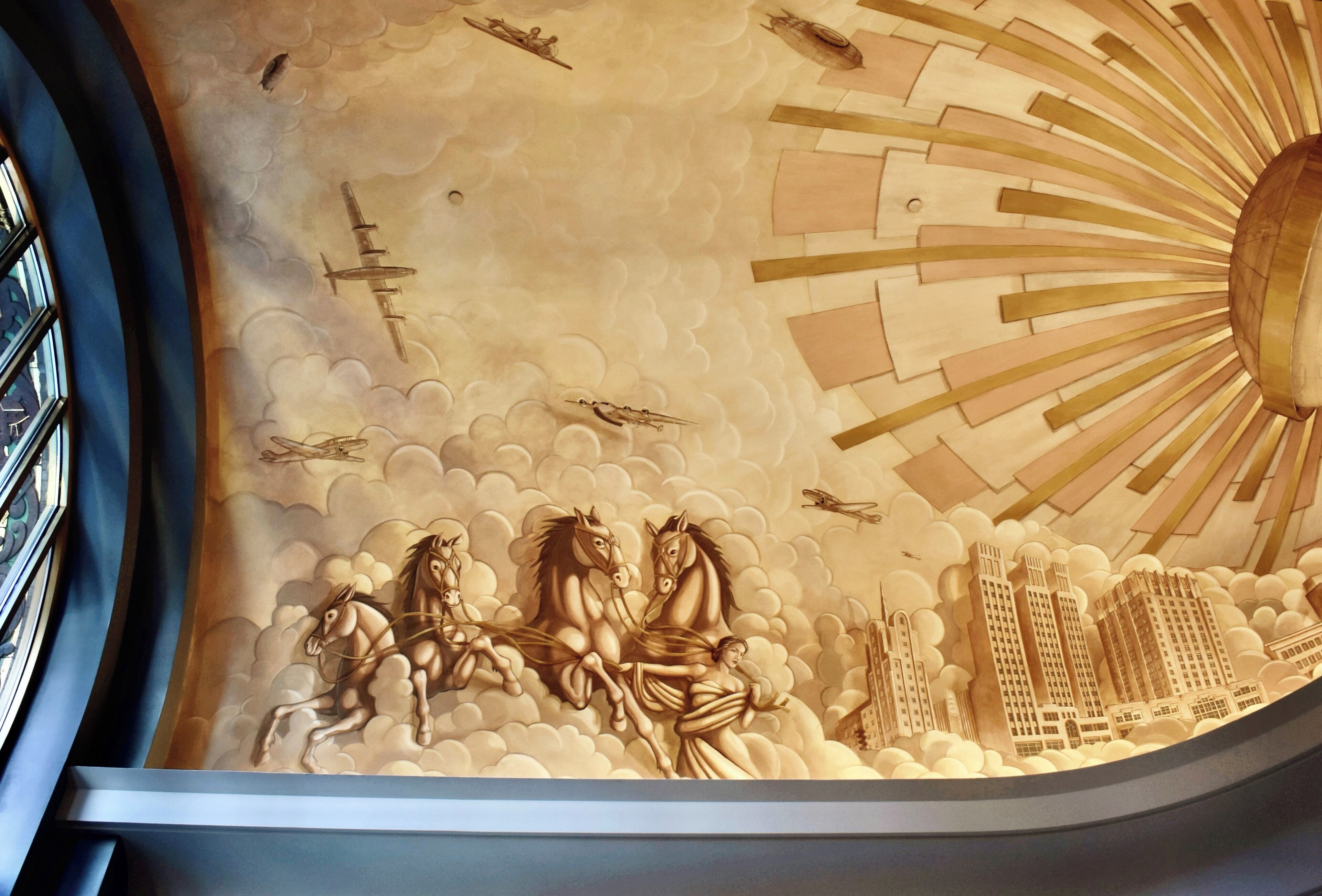 A ceiling mural with a sun, clouds, horses, and buildings.