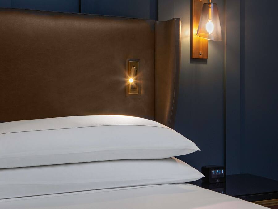 Cama bem arrumada com almofadas brancas e cabeceira, mesa de cabeceira com relógio e luminária fixada na parede.