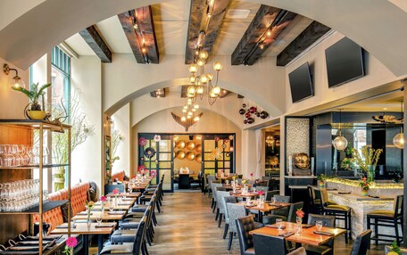 A restaurant with a long dining area, high ceilings, and exposed beams. Tables are set with glassware and centerpieces.
