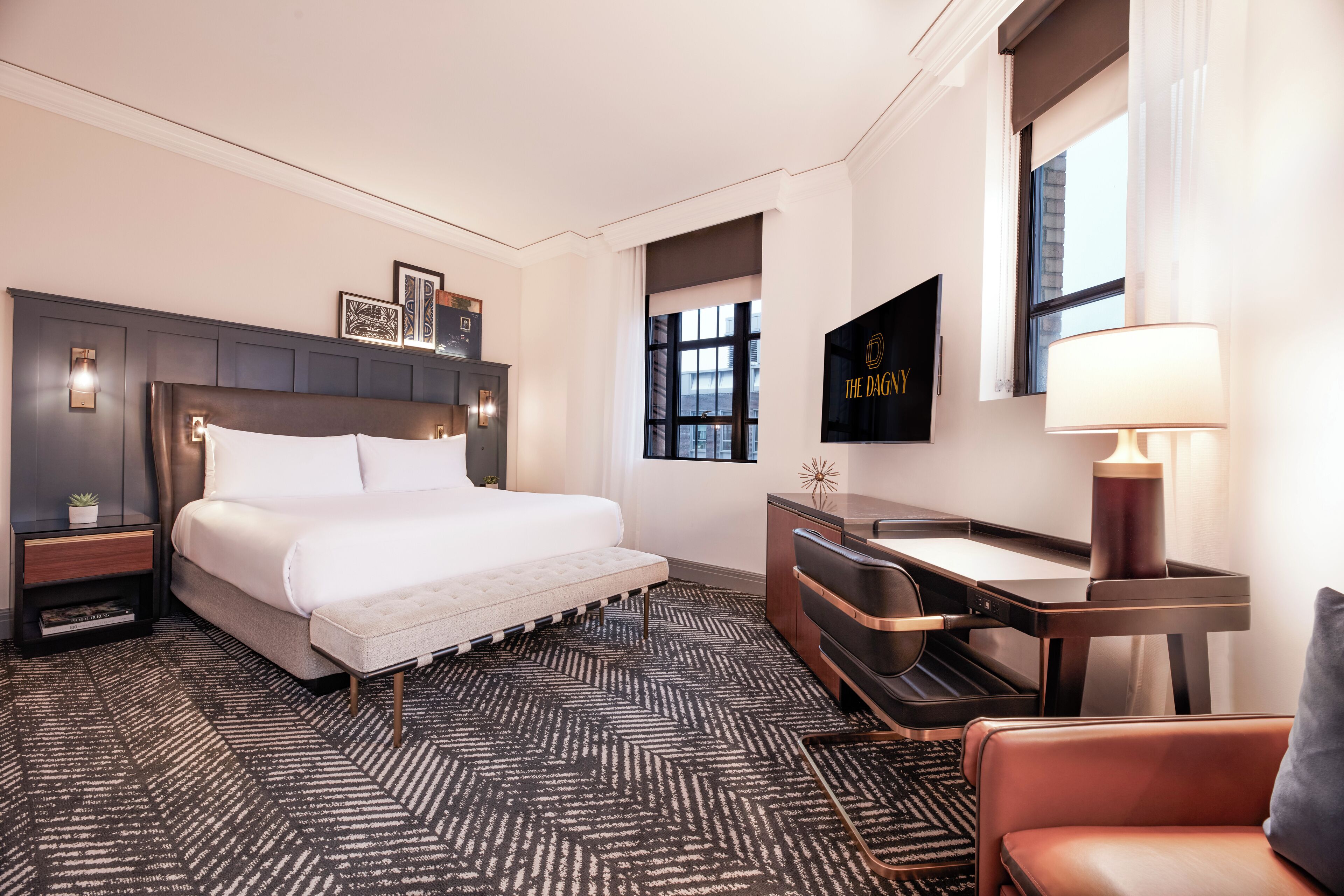 A modern hotel room with a large bed, a desk, and a chair. There is a window with a view of the city, a wall-mounted TV, and a lamp on the desk.