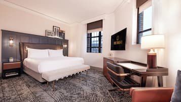 A modern hotel room with a large bed, a desk, and a chair. There is a window with a view of the city, a wall-mounted TV, and a lamp on the desk.