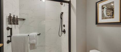 Ducha con cerramiento de vidrio, pared de mármol blanco, una obra de arte enmarcada y un inodoro blanco.