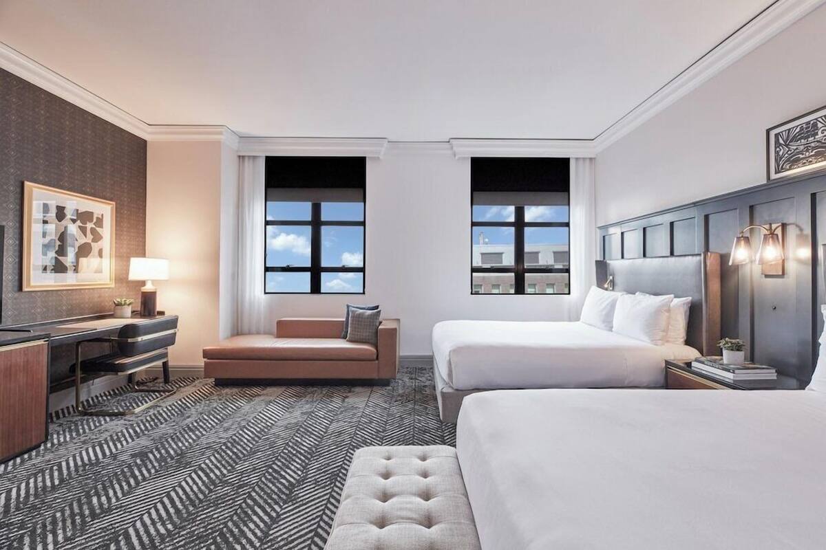 Habitación de hotel con una cama grande, un sofá más pequeño, un escritorio con lámpara y vistas a la ciudad a través de amplias ventanas.