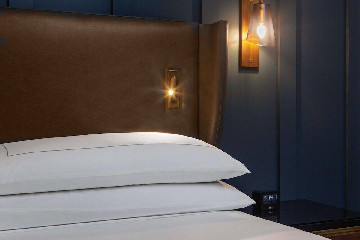Una cama con almohadas blancas y un cabecero, un reloj de noche y una lámpara de pared.