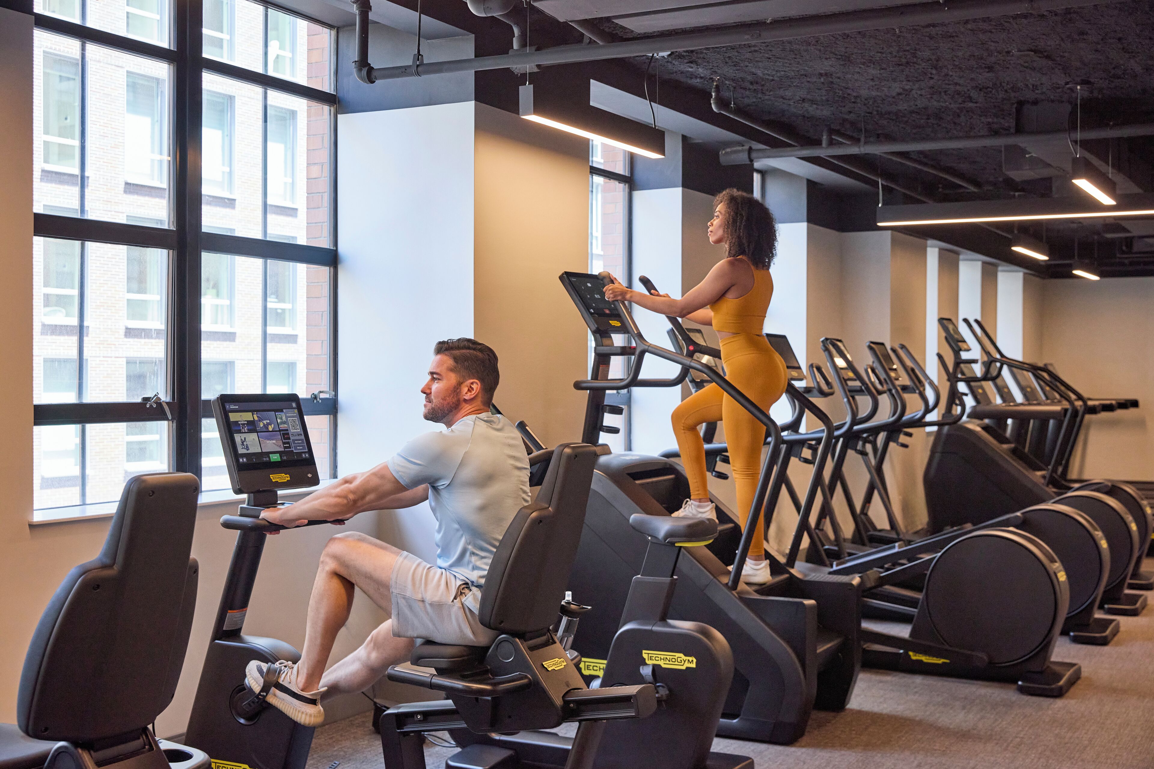 Une salle de sport équipée de tapis de course, où un homme et une femme font de l’exercice, et de grandes fenêtres qui laissent entrer la lumière naturelle.