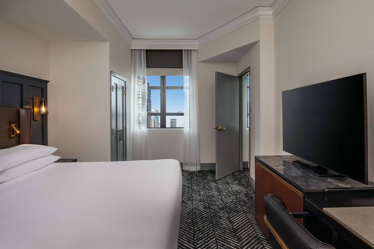 Una habitación de hotel con cama, escritorio con televisor de pantalla plana, vista de la ciudad por una ventana y alfombra estampada.