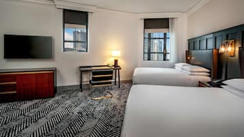 Habitación de hotel con dos camas, un escritorio con lámpara, televisor montado en la pared y vistas a la ciudad a través de las ventanas.
