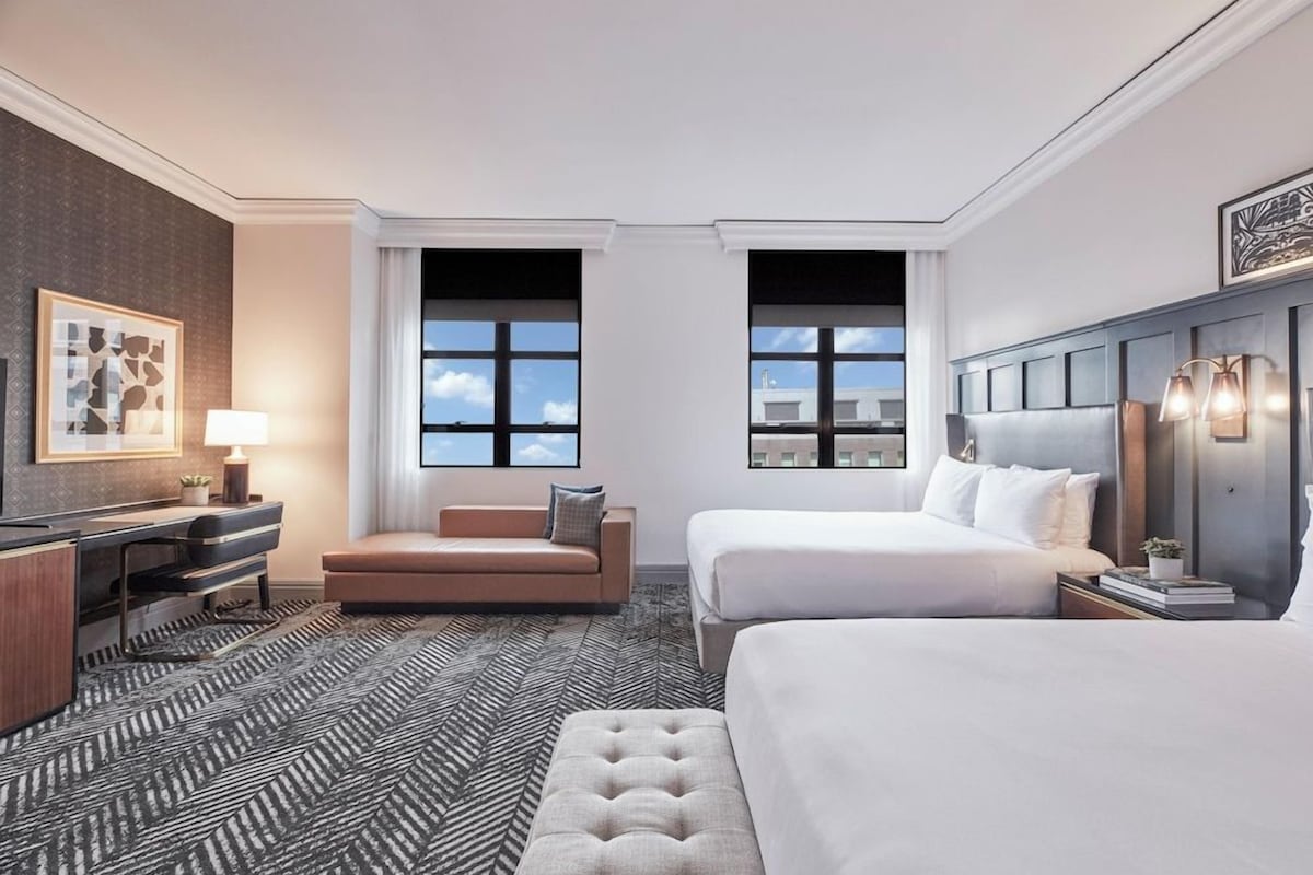 Habitación de hotel con una cama grande, un sofá más pequeño, un escritorio con lámpara y vistas a la ciudad a través de amplias ventanas.