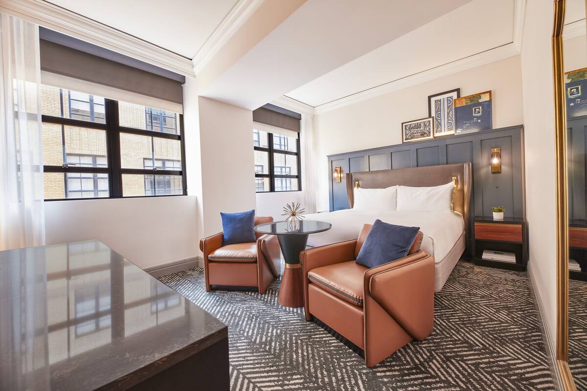 Habitación de hotel con una cama grande, dos sillones de cuero, una mesita y vistas a la ciudad a través de amplias ventanas.