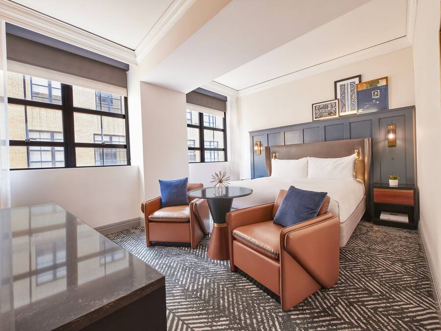 Une chambre d’hôtel avec un grand lit, deux fauteuils en cuir, une petite table et une vue sur la ville grâce à de grandes fenêtres.