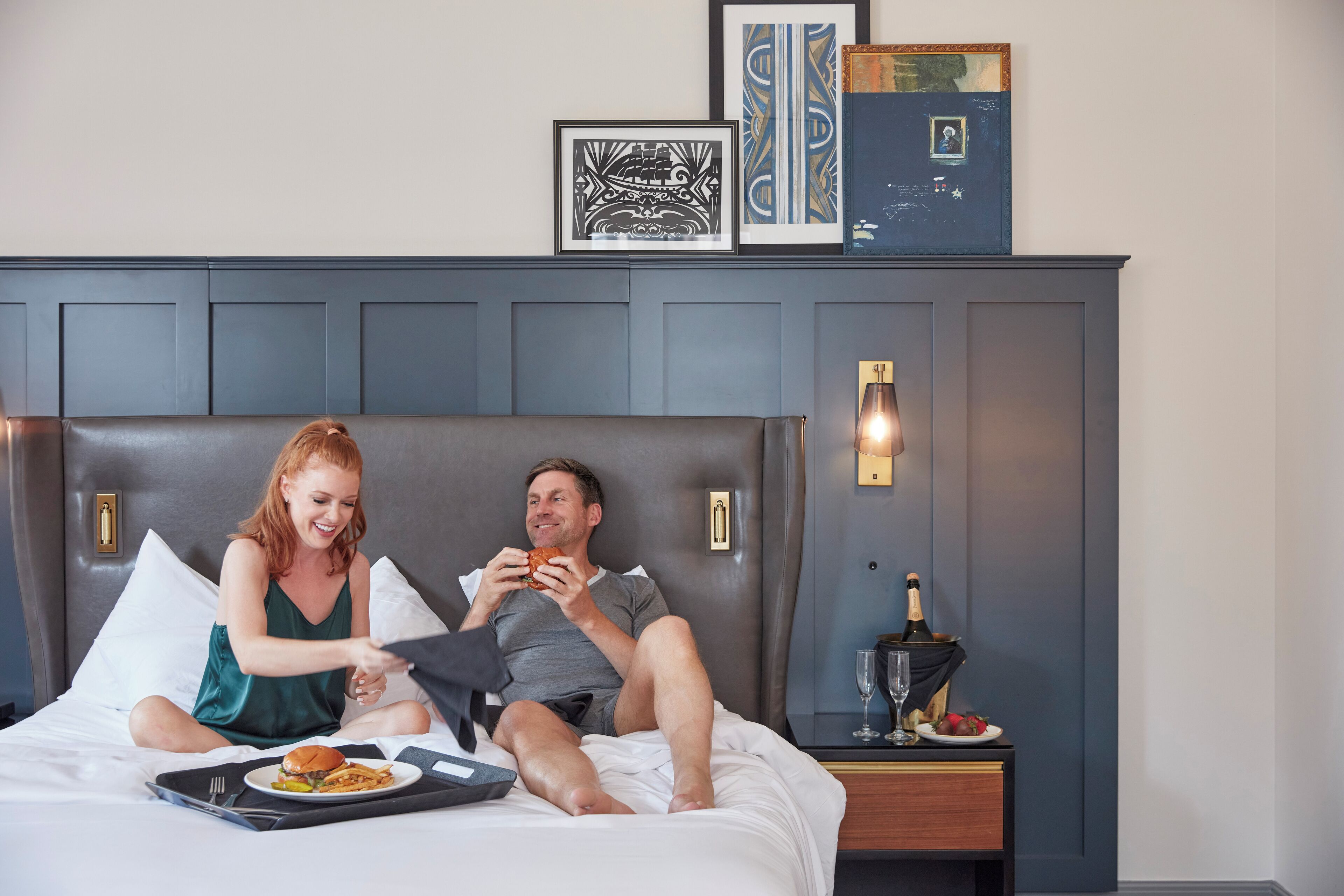 A hotel room with a large bed, a bedside table with a bottle of champagne and glasses, and framed artwork on the wall.
