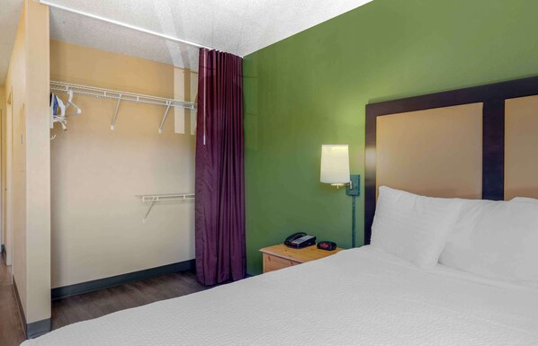 A hotel room with a bed, a nightstand with a lamp, a telephone, and a coat rack with hanging items. - Extended Stay America Suites Boston Waltham 52 4th Ave (Waltham)