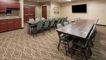 A conference room with a long table and chairs, a television on the wall, and a kitchenette area.