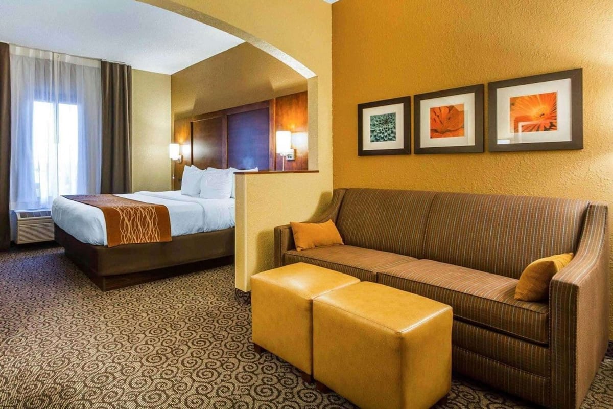 A hotel room with a bed, a sofa, an ottoman, a window with curtains, and three framed pictures on the wall.