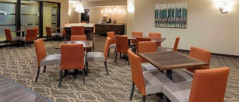 A hotel dining area with multiple tables and chairs, a kitchen in the background, and a large window with frosted glass panels.