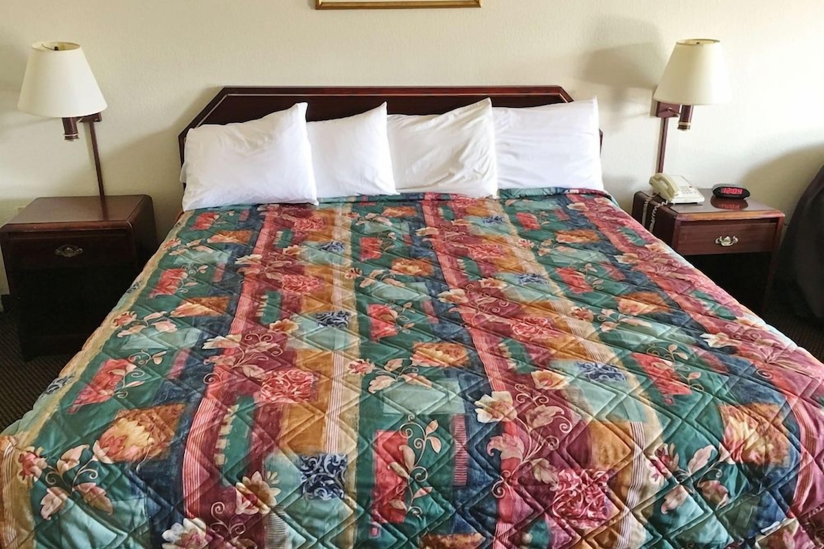 A hotel room with a bed, two bedside tables, a nightstand, a lamp, a clock, and a framed picture on the wall.