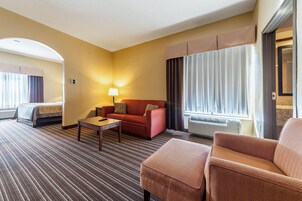 Habitación de hotel con cama, sofá, mesa de centro y una silla. Se ve una ventana con cortinas y un equipo de aire acondicionado.