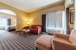 Habitación de hotel con cama, sofá, mesa de centro y una silla. Se ve una ventana con cortinas y un equipo de aire acondicionado.