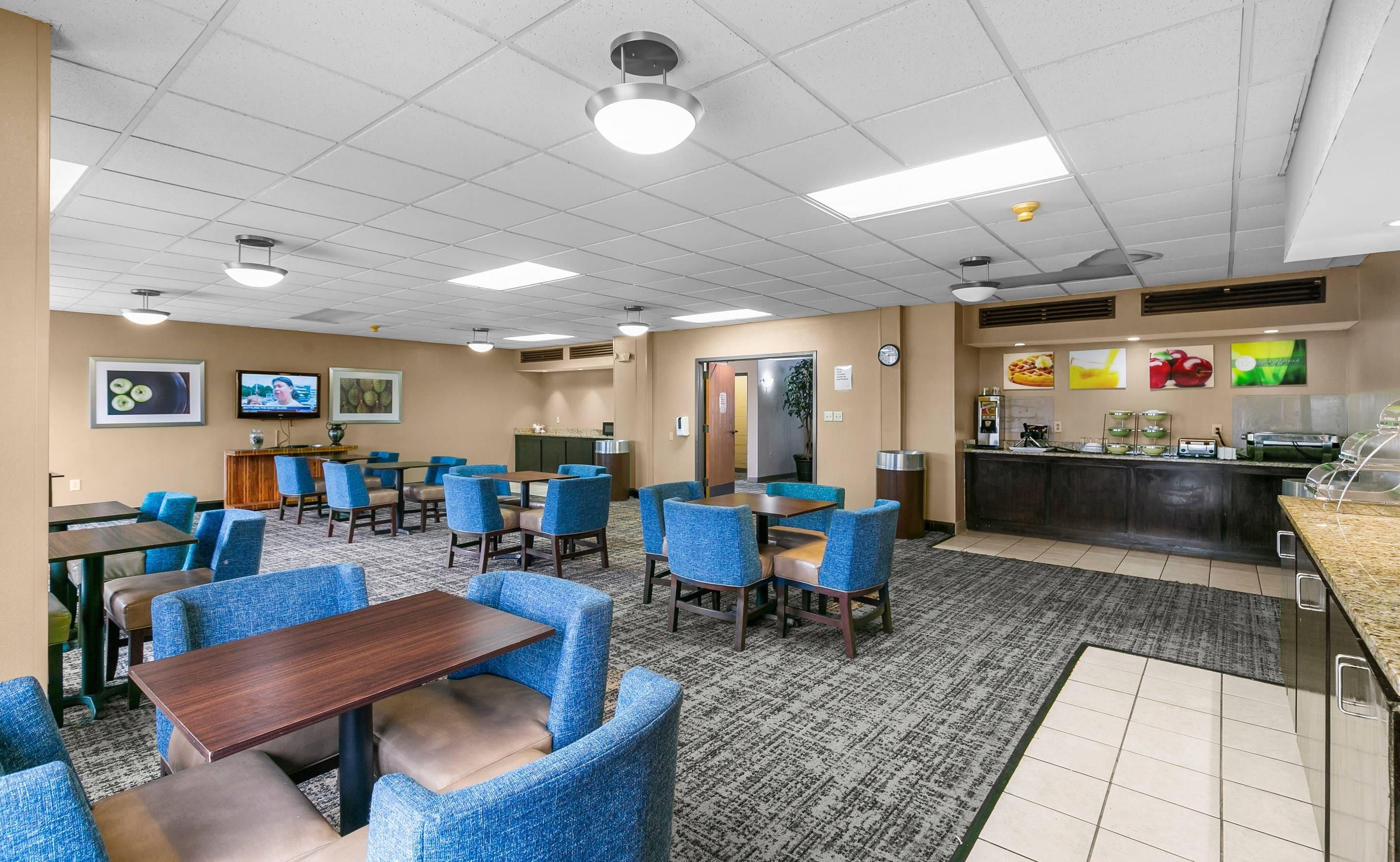 A hotel dining area with multiple tables and chairs, a buffet counter, and a television screen mounted on the wall.