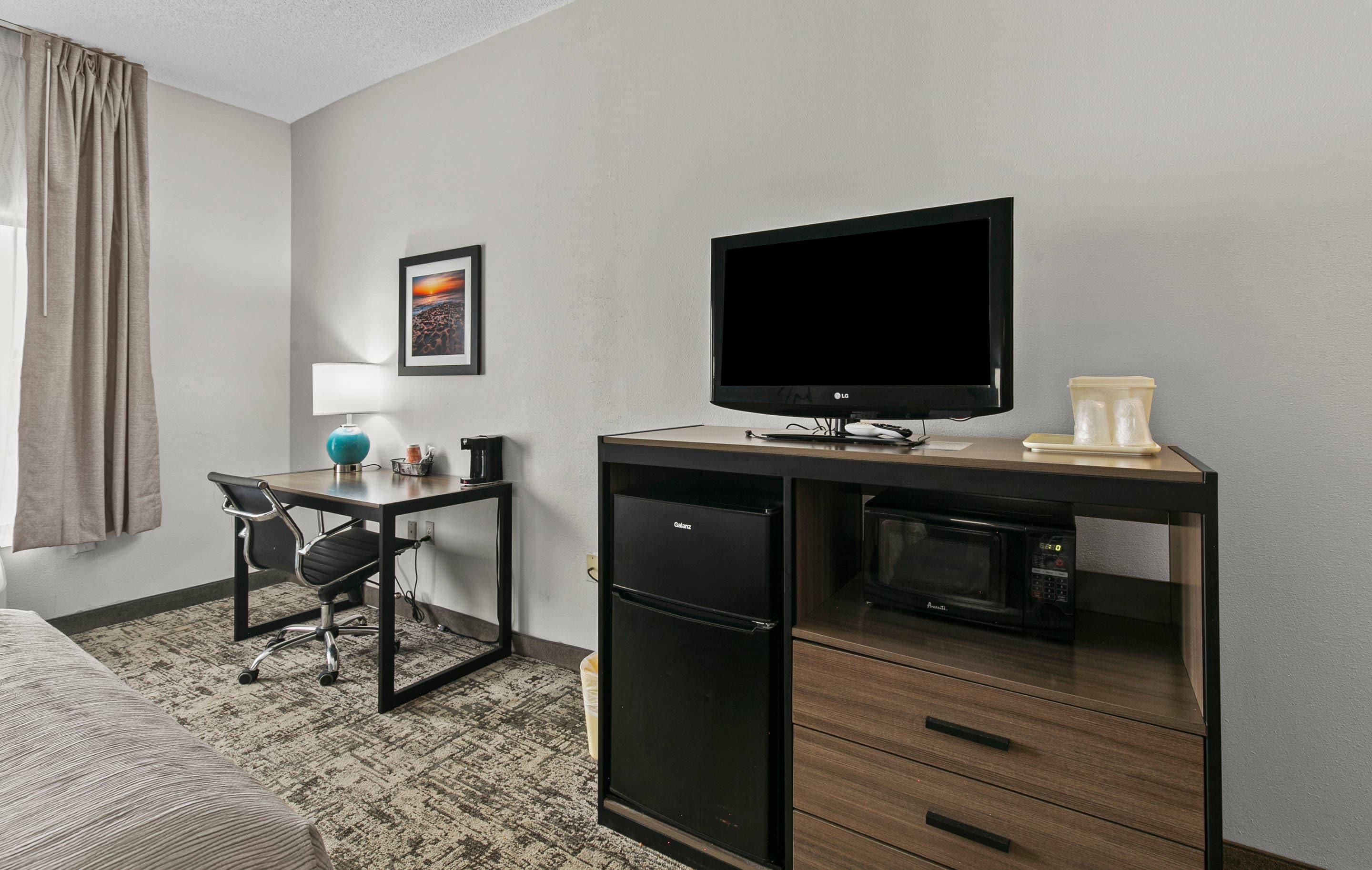 A hotel room with a desk, chair, television, microwave, refrigerator, and a bed.
