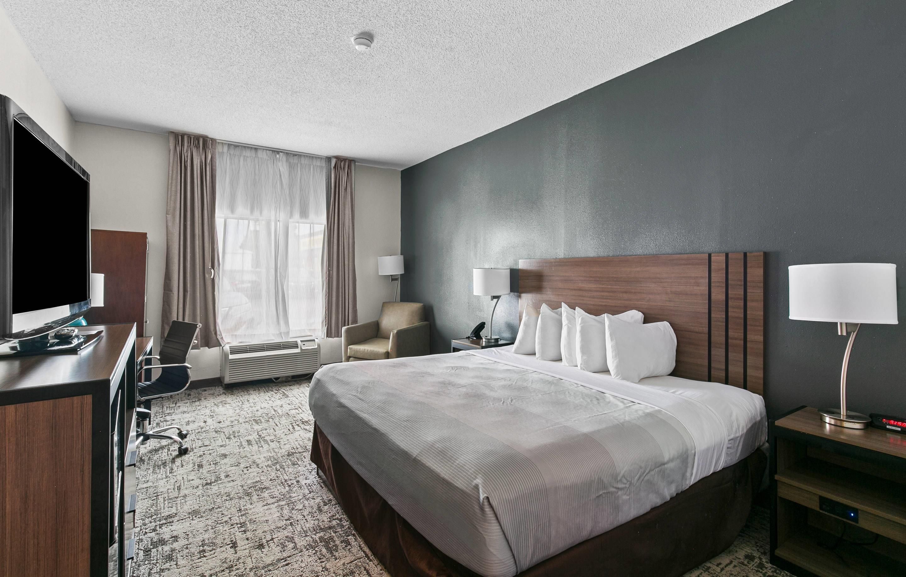 A hotel room with a large bed, a desk with a flat-screen TV, a chair, a desk lamp, and a bedside table with a lamp.