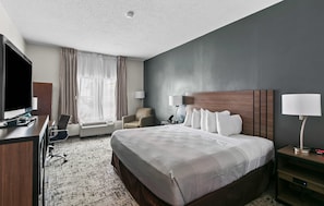 Una habitación de hotel con una cama grande, un escritorio con televisor de pantalla plana, una silla, una lámpara de escritorio y una mesita de noche con lámpara.