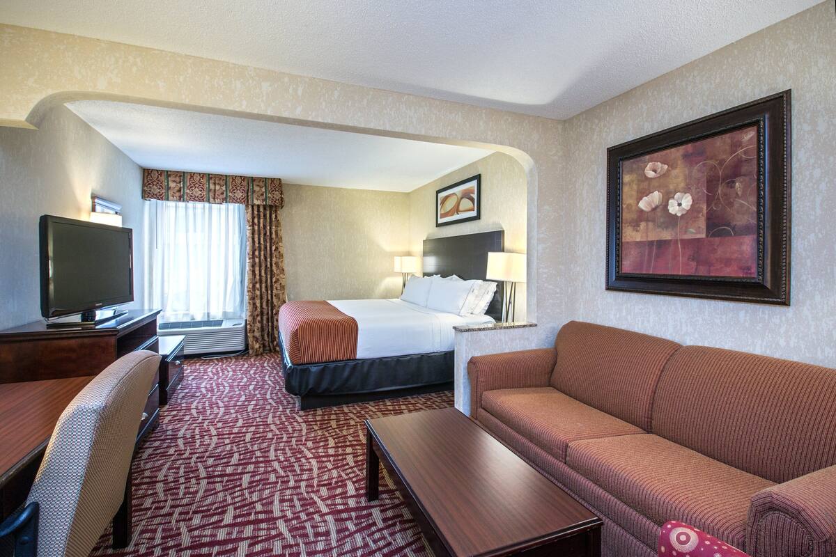 A hotel room with a bed, a sofa, a desk, a TV, and a painting on the wall.