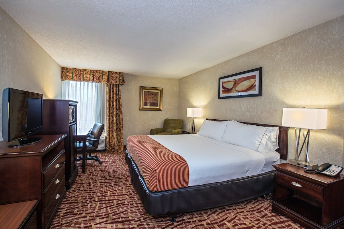 A hotel room with a bed, a desk with a television, a chair, a lamp, and a framed picture on the wall.