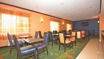 A hotel dining area with multiple tables and chairs, a flat-screen TV mounted on the wall, and a carpeted floor with a floral pattern.