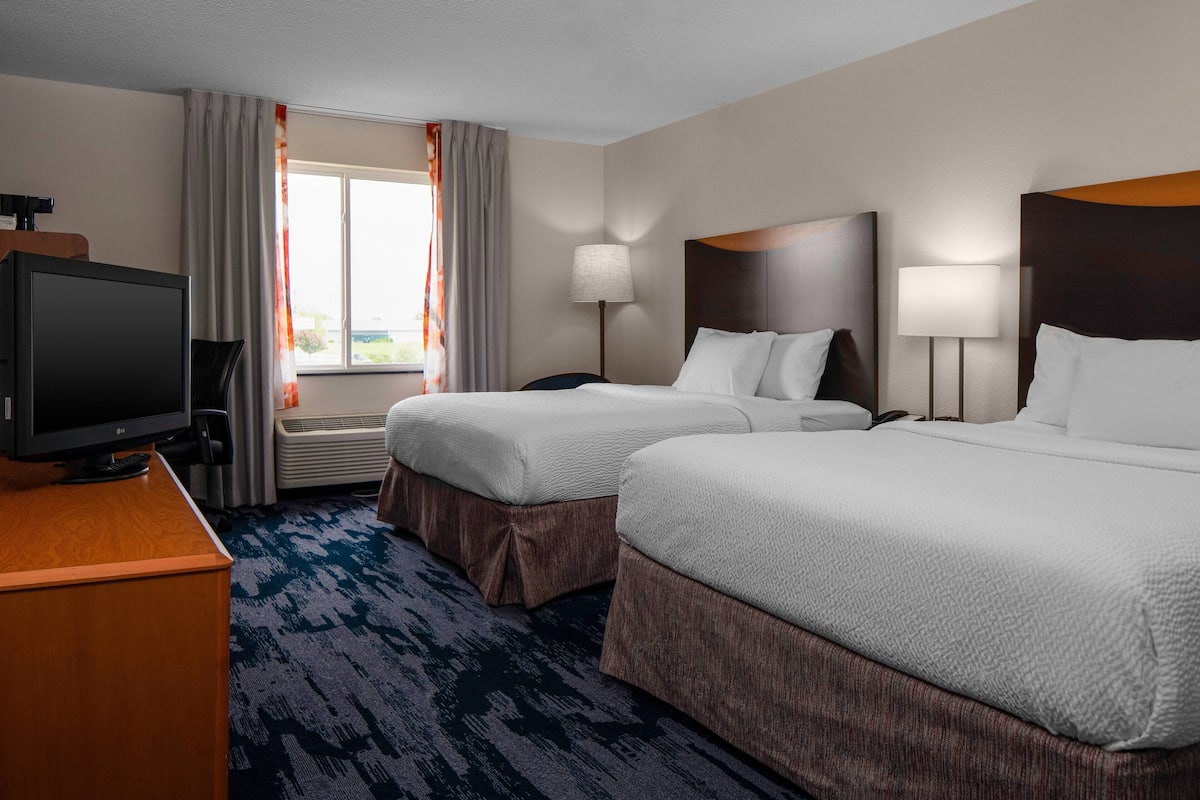 Ein Hotelzimmer mit zwei Betten, einem Fernseher, einem Schreibtisch und einem Fenster mit Vorhängen.