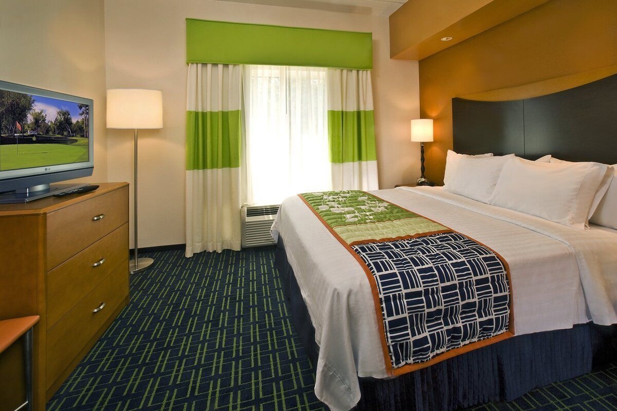 A hotel room with a bed, a television on a dresser, a lamp, and a window with curtains.