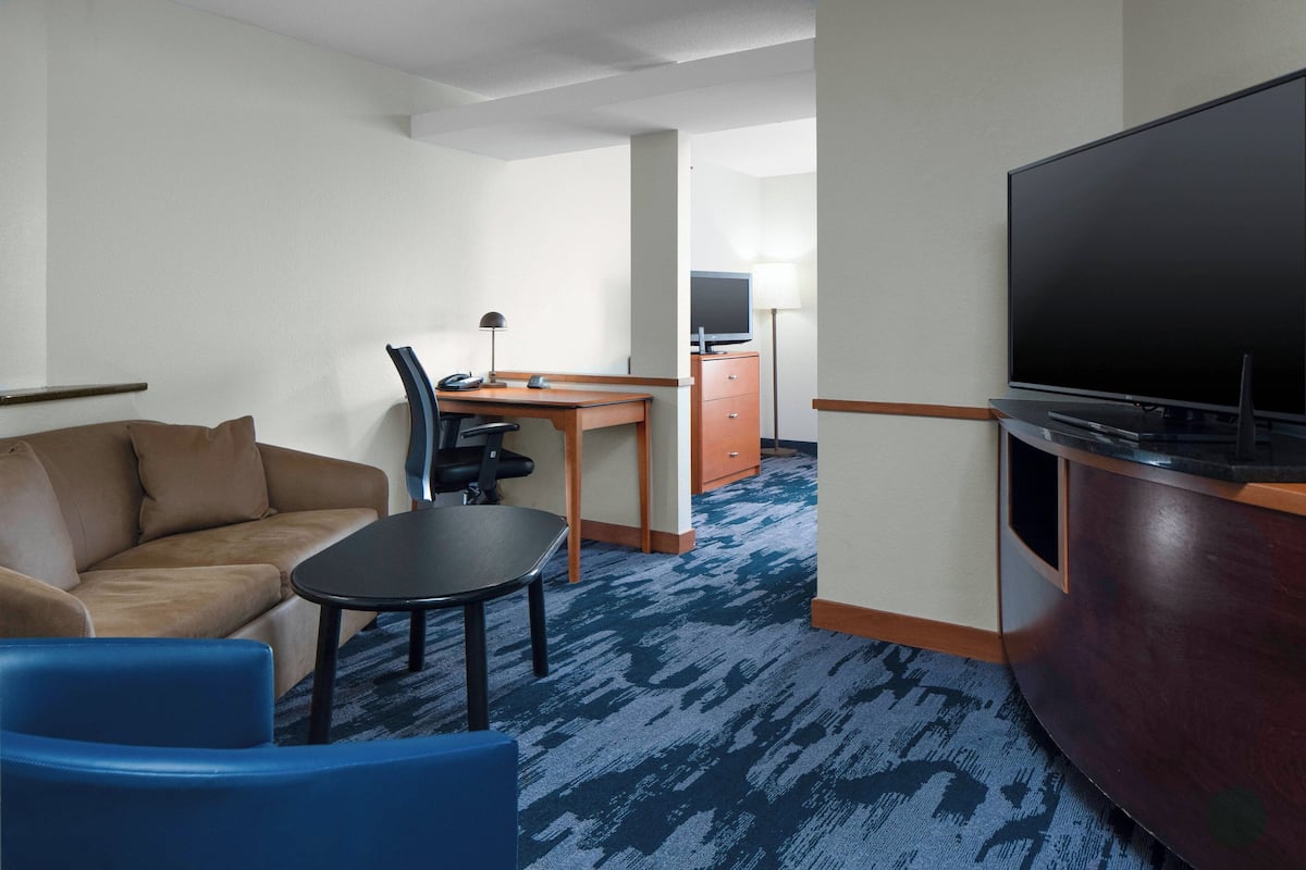 Ein Hotelzimmer mit einem braunen Sofa, einem blauen Sessel, einem kleinen Tisch, einem Schreibtisch mit Stuhl, einem Fernseher und einem Schrank.