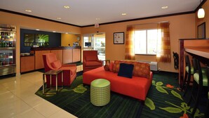 A hotel room with a red sofa, a small table, a green ottoman, a kitchenette, and a dining area with chairs. - Fairfield Inn & Suites by Marriott - Jefferson City (Jefferson City)