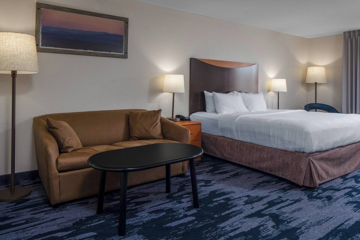 Une chambre d’hôtel avec un canapé marron, une petite table ronde, un lit avec une literie blanche, deux lampes de chevet et un tableau au mur.