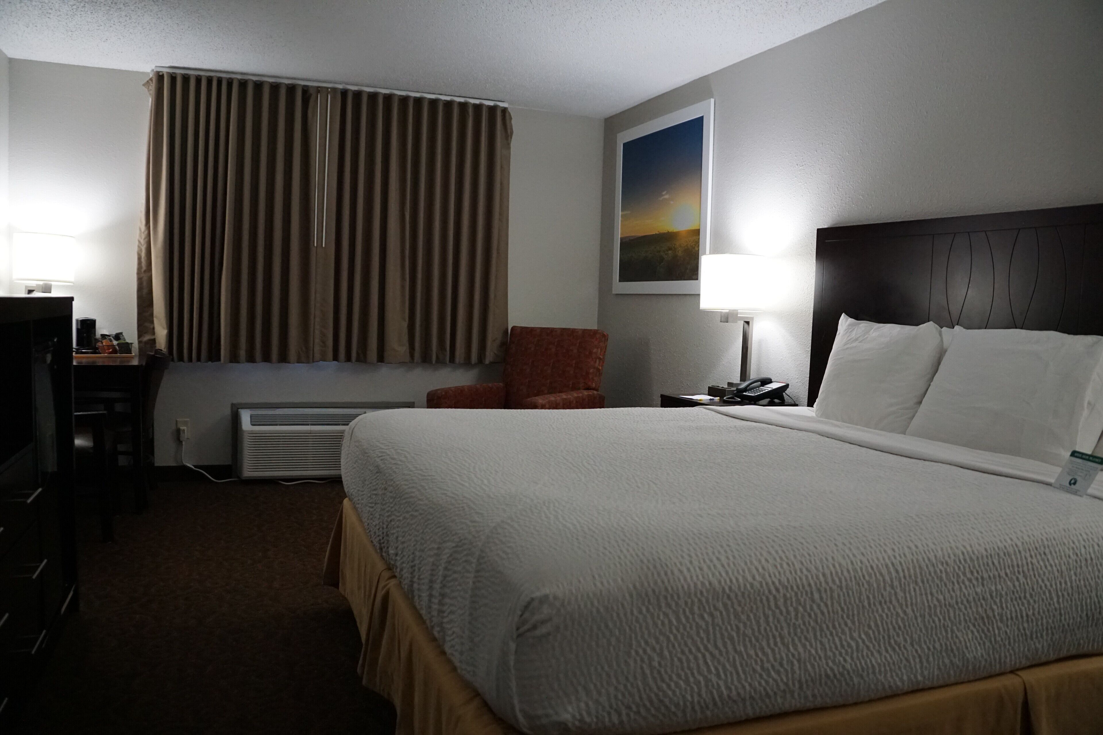 Quarto de hotel com uma cama grande, uma mesa com cadeira, uma janela com cortinas, uma mesa de cabeceira com abajur e um quadro na parede.