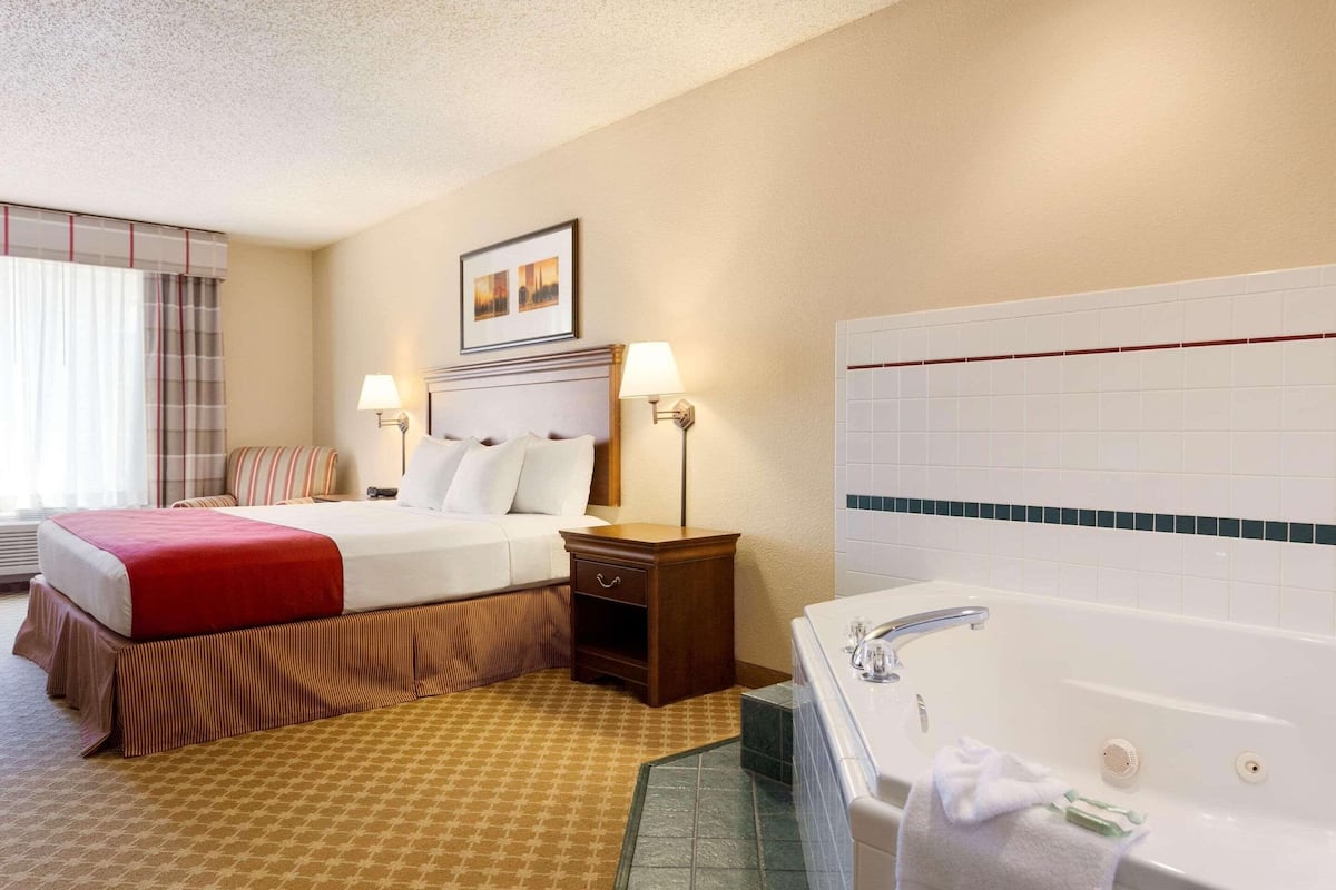 A hotel room with a large bed, a bedside table, a lamp, a bathroom with a bathtub, and a tiled wall.