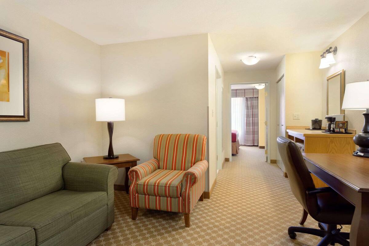 A hotel room with a green sofa, striped armchair, desk with a lamp, and a mirror.