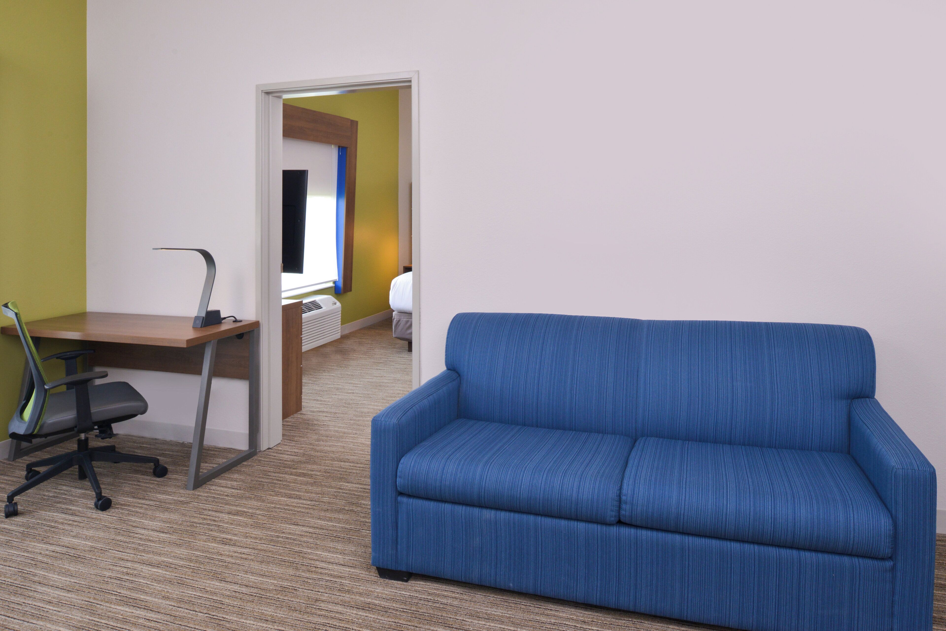 A room with a blue sofa, a wooden desk with a chair, and a bed in the background.