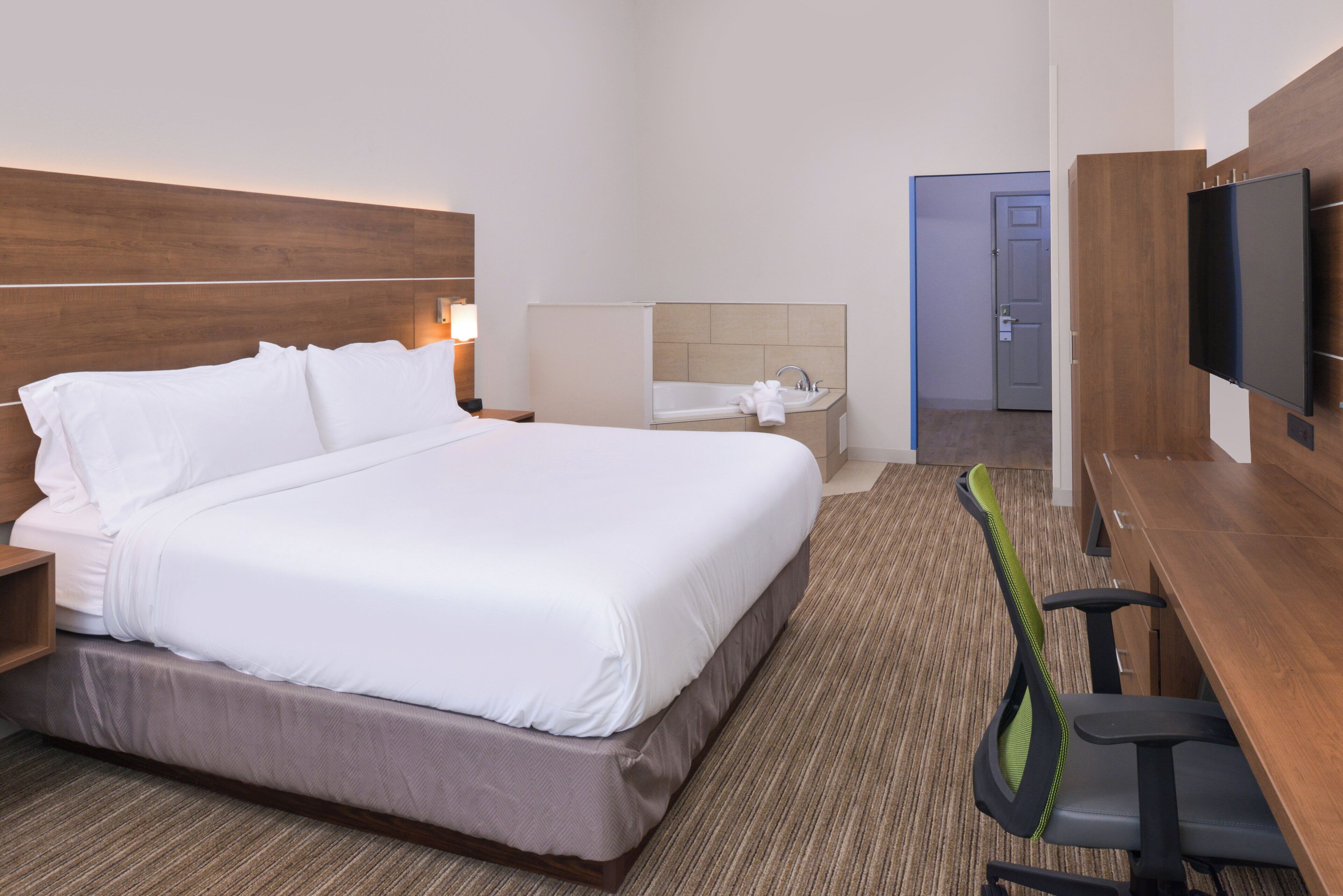 A hotel room with a large bed, a desk, a chair, a television, a bathroom with a bathtub, and a door leading to another room.