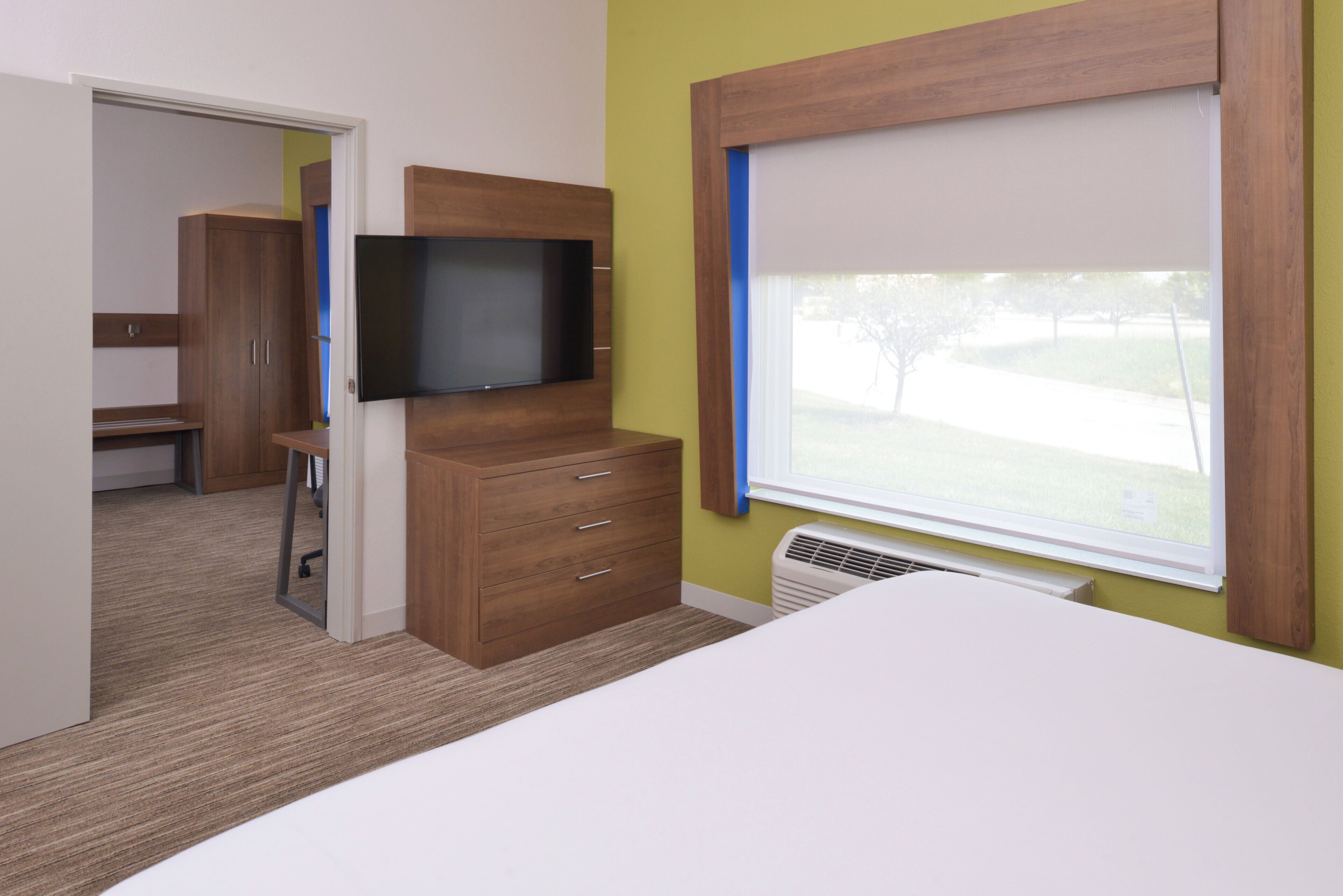 Une chambre d’hôtel avec un lit, un téléviseur fixé sur une armoire en bois, une fenêtre avec des stores et une porte donnant sur une autre pièce.