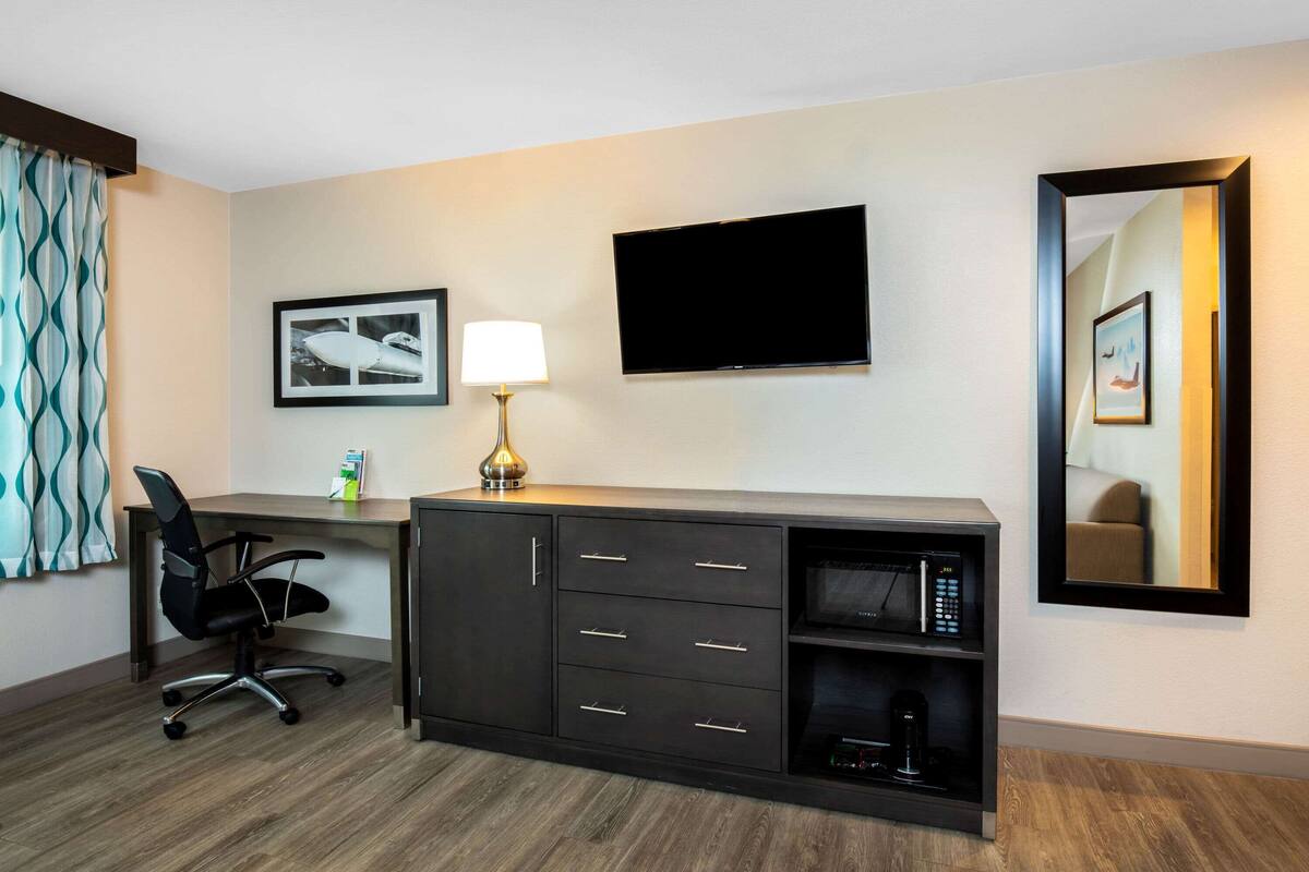 A hotel room with a desk, a chair, a flat-screen TV, a lamp, a framed picture, and a mirror.