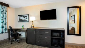 A hotel room with a desk, a chair, a flat-screen TV, a lamp, a framed picture, and a mirror.