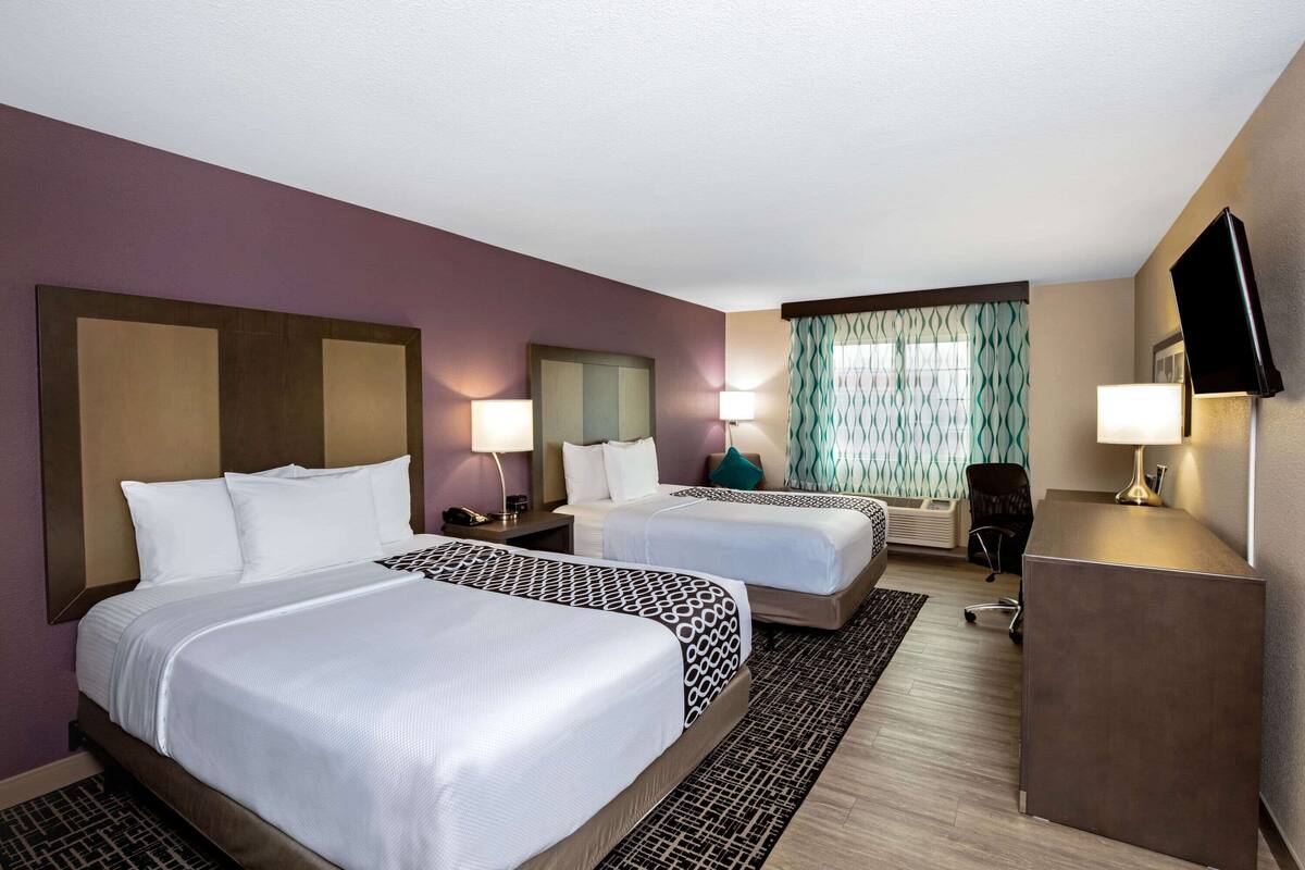 A hotel room with two beds, a desk, a TV, and a window with curtains.