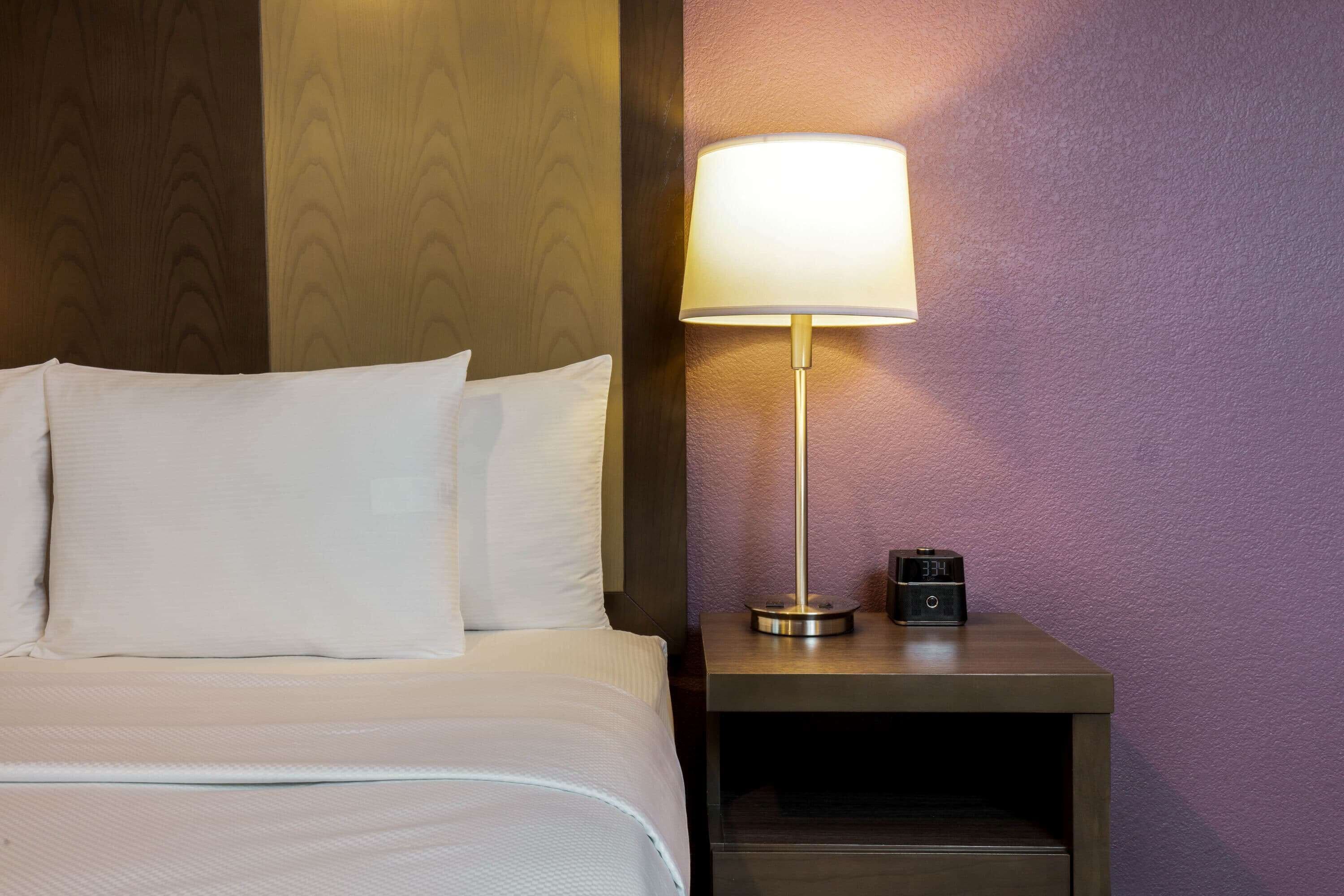 A neatly made bed with white linens, a bedside lamp, and a small clock on a wooden nightstand.