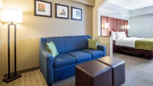 A hotel room with a blue sofa, a brown ottoman, a bed with a green blanket, and framed pictures on the wall. - Comfort Suites at Woodbridge (Avenel)