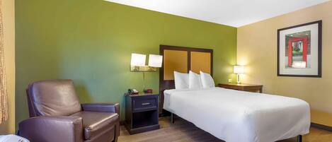 A hotel room with a bed, a chair, a nightstand, a lamp, and a framed picture on the wall.