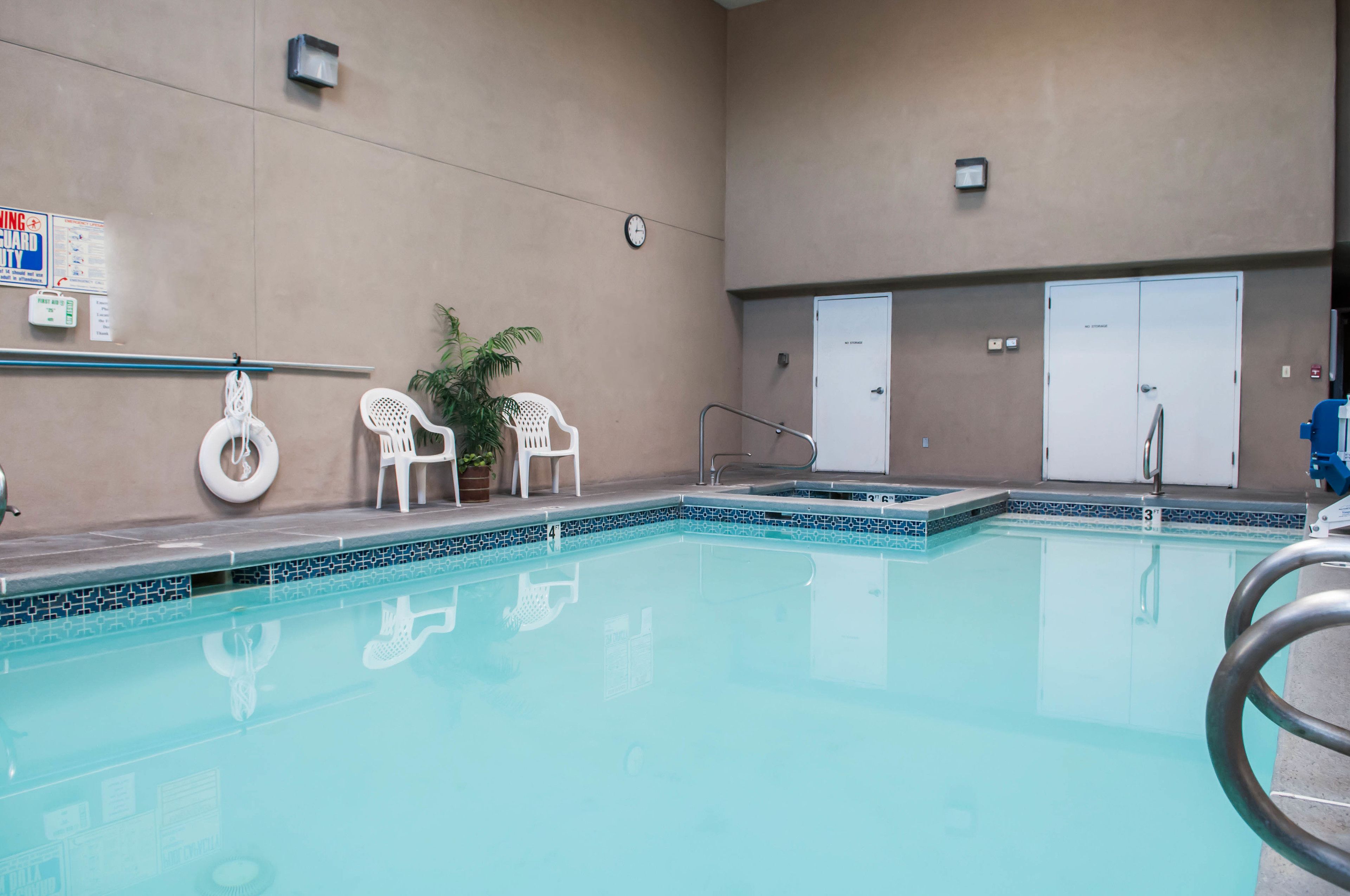 A swimming pool with a tiled edge, two white chairs, a plant, and a lifeguard sign on the wall.