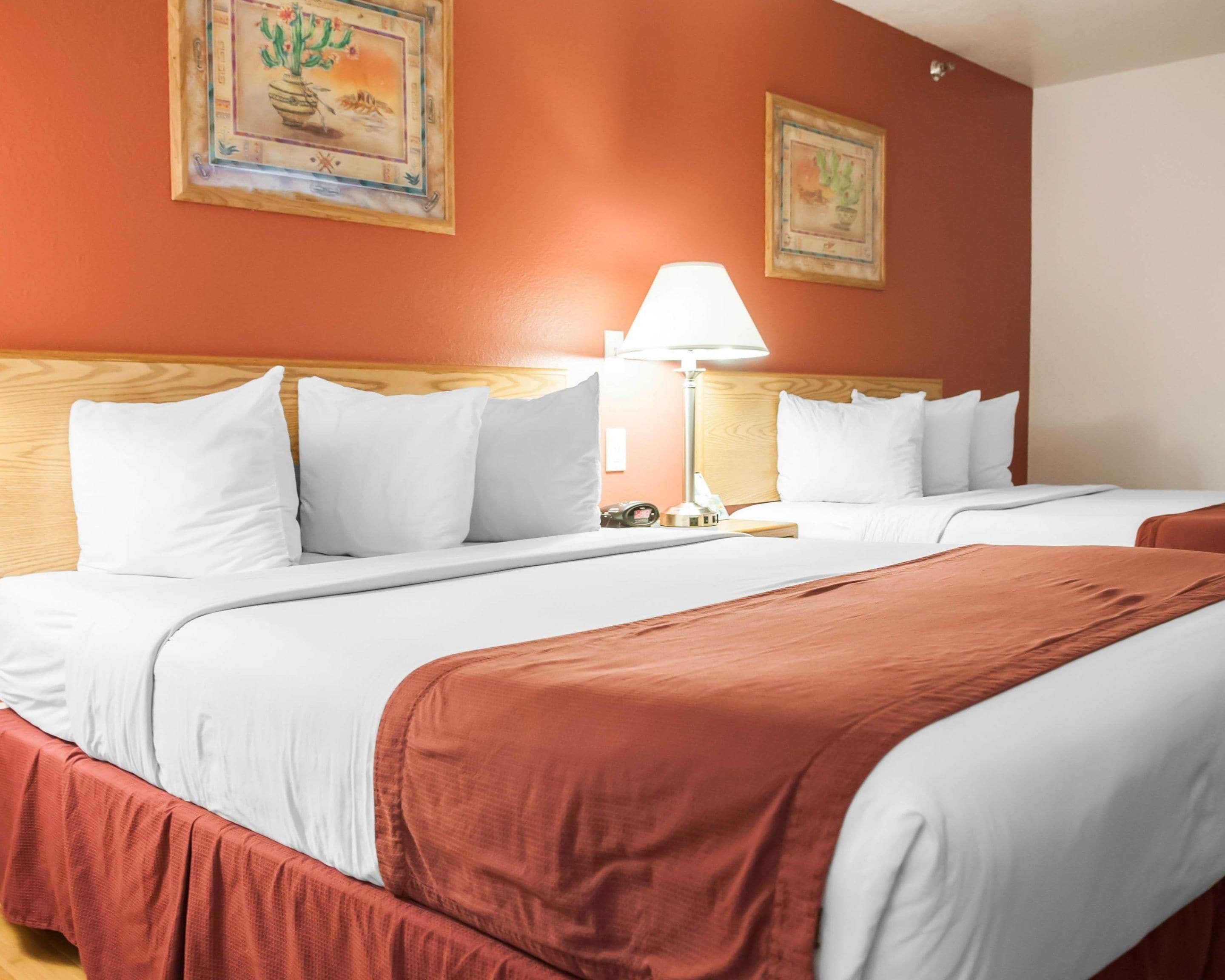 Habitación de hotel con dos camas, ropa de cama blanca, cabecero de madera, mesita de noche con lámpara y dos cuadros enmarcados en la pared.