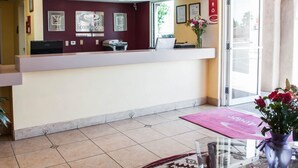 Un mostrador de recepción con una computadora, una impresora y un jarrón con flores. En la pared hay certificados enmarcados, y se ve una señal de salida.