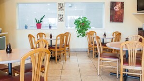 Un comedor con mesas y sillas de madera, plantas en macetas y un ventanal con persianas.